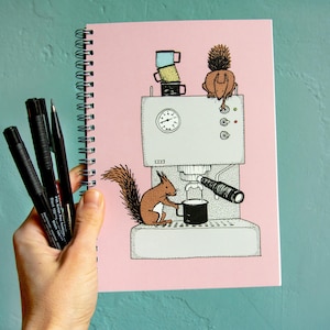 Könnte beinhalten: Ein rosa Notizbuch mit Spiralbindung und einer Schwarzweißillustration von zwei Eichhörnchen, die eine Kaffeemaschine benutzen. Die Illustration zeigt ein Eichhörnchen, das eine Tasse mit Kaffee füllt, und das andere Eichhörnchen steht auf der Maschine.