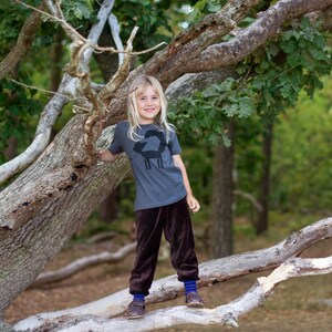 May include: A young person wearing a gray t-shirt with a black fox graphic and brown pants stands on a tree branch in a forest. The t-shirt has the text "Fox" on it.