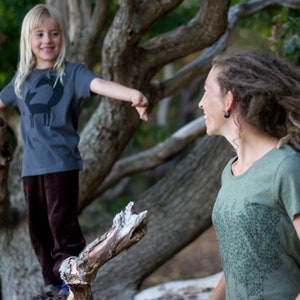 May include: A young girl wearing a gray t-shirt with a black fox graphic stands on a tree branch. She is looking at a woman with long brown dreadlocks wearing a green t-shirt with a tree graphic. The woman is smiling.