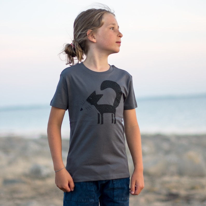 May include: A child wearing a charcoal grey t-shirt with a black silhouette of a fox on the front. The fox is facing left and has a long tail.