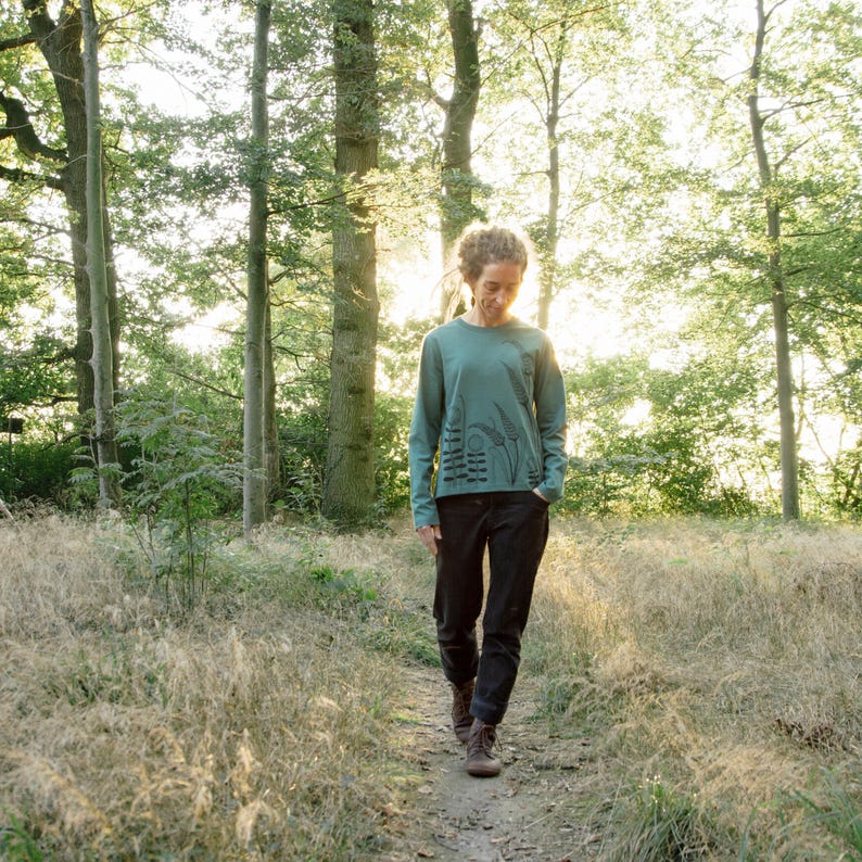 May include: A person wearing a teal long-sleeved shirt with a black graphic print of a forest scene and the word "Love" on it. The person is walking on a path in a forest.