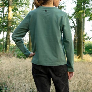 May include: A person wearing a long-sleeved, sage green t-shirt with a small logo on the back. They are wearing black jeans and standing in a field of long grass.