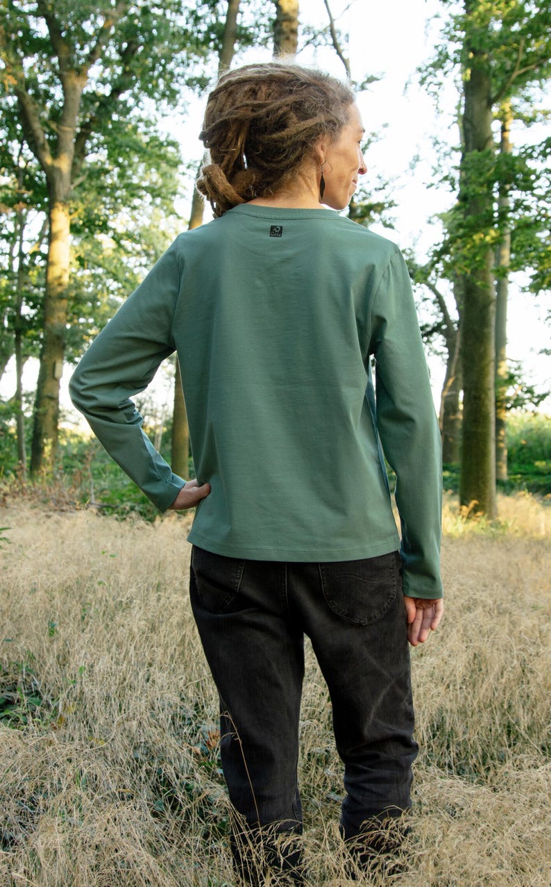 May include: A person wearing a long-sleeved, sage green t-shirt with a small logo on the back. They are wearing black jeans and standing in a field of long grass.
