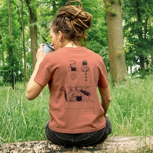 May include: A rust-colored t-shirt with a black graphic of a coffee machine, a squirrel, and other elements. The person is sitting on a log in a grassy area, holding a mug.