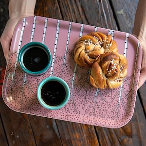 Könnte beinhalten: Ein rosa Serviertablett mit Birkenmuster hält zwei kleine türkisfarbene Kaffeetassen und zwei Zimtschnecken. Das Tablett ist rechteckig und hat ein dekoratives Muster aus weißen Birken und kleinen Punkten.
