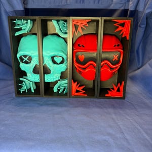 Peut inclure: Une œuvre d'art encadrée en noir, divisée en quatre sections. Les deux premières sections présentent des crânes turquoise avec des motifs d'yeux variés. Les deux dernières sections affichent des casques rouges avec des détails noirs et des motifs de cœur et de X. Le fond est bleu.