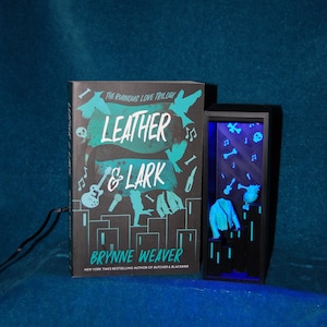 Könnte beinhalten: Ein Buch mit dem Titel "Leather & Lark" von Brynne Weaver, mit türkisfarbenem und weißem Text und Grafiken. Daneben eine schwarze Vitrine mit blauem Leuchten, mit einer Jacke und anderen Objekten.