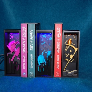 Könnte beinhalten: Ein Satz dekorativer Buchstützen mit beleuchteten Szenen. Die Buchstützen zeigen Titel wie "Butcher & Blackbird", "Leather & Lark" und "Scythe & Sparrow". Jede Buchstütze hat ein einzigartiges Design mit einem Vogel, einer Figur und einer Sense. Die Buchstützen sind schwarz mit farbigen Akzenten.