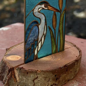 May include: A stained glass panel featuring a heron, set in a wooden base. The heron is depicted in shades of blue, white, and gray against a teal background. The panel also includes cattails in shades of brown and green. The wooden base is a cross-section of a tree trunk.