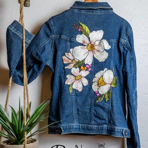 May include: A blue denim jacket featuring embroidered white and pink floral designs with green leaves. The jacket is suspended on a wooden hanger. The text "Re New Upcycled Goods" is visible at the bottom.