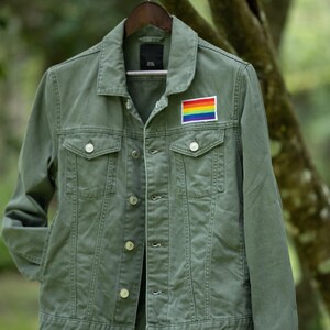 May include: An olive green denim jacket with a rainbow flag patch. The jacket features button fastenings, two chest pockets, and a traditional collar. It hangs on a wooden hanger against a blurred green backdrop.