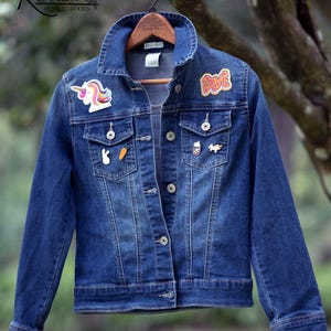 Puede incluir: Chaqueta vaquera azul con varios parches, incluyendo un unicornio, la palabra "Love", una zanahoria y un té boba. La chaqueta está colgada en una percha de madera sobre un fondo verde borroso.