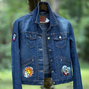 May include: A blue denim jacket with patches of a sun, a dog, and other designs. The jacket has a classic collar, button closures, and pockets. The text "Re New Upcycled Goods" is visible in the background.