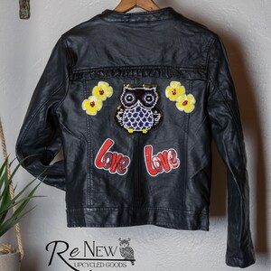 May include: Black leather jacket with an owl and flower patches. The back of the jacket features a sequined owl, yellow flower patches, and the word "Love" in red. The jacket is hanging on a wooden hanger.