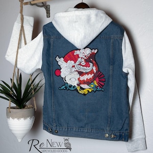 May include: A denim jacket with a white hooded sweatshirt attached. The back of the jacket features a colorful koi fish patch. The jacket is hanging on a wooden rod with a plant in a hanging pot.
