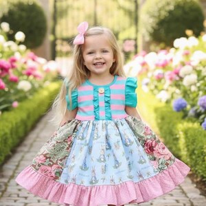 Può includere: Una bambina indossa un colorato vestito di Pasqua con un fiocco rosa tra i capelli. L'abito presenta un corpetto a righe blu e rosa, una gonna con stampa floreale e coniglietti e un bordo rosa a pois. Indossa scarpe bianche.