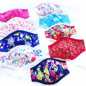 May include: A collection of handmade face masks in various floral and patterned fabrics. The masks feature designs in pink, blue, red, and white, with elastic ear loops for a comfortable fit. The masks are made of fabric.