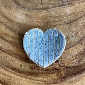 Op de afbeelding: Een hartvormig keramisch schaaltje met een lichtblauwe glazuur en verticale, getextureerde lijnen. Het schaaltje staat op een houten achtergrond, wat de handgemaakte kwaliteit en het unieke ontwerp benadrukt. Dit decoratieve stuk is ideaal voor het bewaren van kleine items.