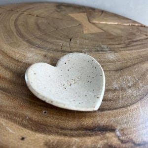 Op de afbeelding: Een klein, hartvormig keramisch schaaltje met een gespikkelde, gebroken witte afwerking. Het woord "xoxo" is subtiel op het oppervlak van het schaaltje gegraveerd. Het schaaltje ligt op een getextureerd, houten oppervlak, wat een rustieke uitstraling creëert.