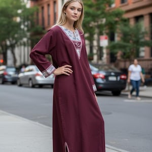 May include: A long, burgundy dress with long sleeves and a detailed neckline and cuffs. The dress has a relaxed fit and a slit on the side. The model is standing on a city street.