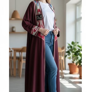 May include: A long, burgundy open-front coat with patterned trim in red, white, and blue. The coat is worn over a white t-shirt and blue jeans. The coat's trim features a geometric design. The outfit is completed with white sneakers.