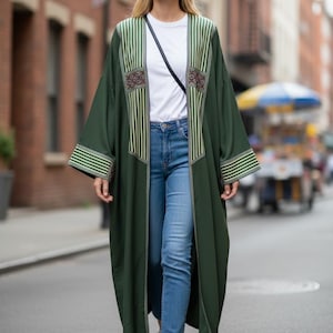 May include: A long, dark green open-front duster coat featuring vertical striped panels on the chest and sleeves, worn over a plain white t-shirt, blue denim jeans, and white sneakers, with a thin black crossbody strap visible across the chest.