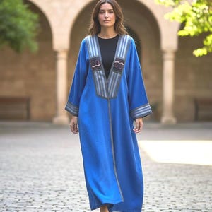 May include: A vibrant blue kaftan featuring a deep V-neckline with vertical grey and white striped panels. The garment includes intricate dark embroidery details on the chest and matching striped cuffs on the long sleeves, worn over a black top.