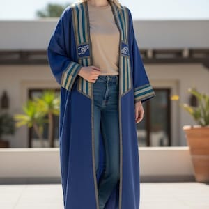 May include: A long, flowing blue robe with decorative gold and teal accents. The robe features vertical stripes and ornate designs on the front panels and sleeves. The model is wearing a tan shirt and blue jeans, with white sneakers.