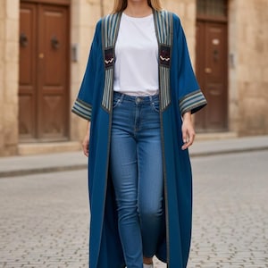 May include: A long, open-front teal blue duster coat featuring vertical striped panels along the lapels and cuffs, worn over a plain white t-shirt and blue denim jeans with white sneakers. The coat has decorative embroidered accents on the chest.