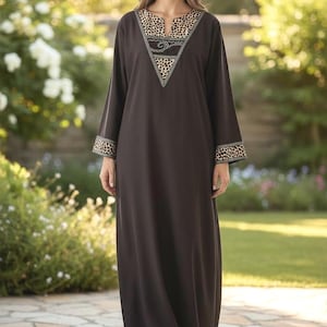 May include: A long, dark brown dress with long sleeves. The neckline and cuffs feature a leopard print design. The dress is worn by a person standing in a garden setting.
