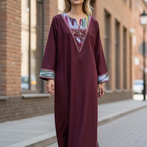 May include: A burgundy-colored, long-sleeved dress with a V-neckline and a floor-length hem. The neckline and sleeve cuffs feature colorful patterned fabric. The dress is worn by a person standing on a brick-paved walkway.