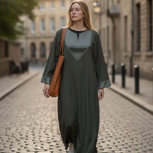 May include: An olive green maxi dress with a black neckline and striped detailing on the bodice and sleeves. The dress is paired with a brown tote bag and white sneakers. The model is walking on a cobblestone street.