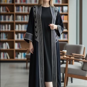 May include: A black open-front abaya featuring silver embroidery along the lapels and sleeve cuffs, worn over a matching black dress. The garment has long, wide sleeves and a floor-length silhouette with vertical silver striped detailing.