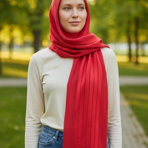 May include: A woman wearing a red hijab and scarf, with a cream-coloured long-sleeve top and blue jeans. The red scarf features vertical stripes. The background is a blurred outdoor scene with green trees.