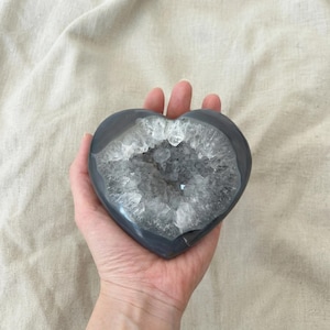 May include: A heart-shaped, gray geode crystal with a sparkling, crystalline interior. The geode is held in a hand, showcasing its polished exterior and natural, textured cavity. The crystal is a unique decorative item.