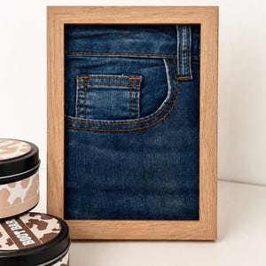 Pode incluir: Uma impressão emoldurada com um close-up de um bolso de jeans azul denim com costura laranja. A moldura é de madeira clara. Duas latas redondas com estampa de vaca e o texto "BOOT BAR" estão em primeiro plano.