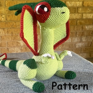 Puede incluir: Peluche de dinosaurio Dilophosaurus tejido a ganchillo en tonos verdes, rojos y blancos. El dinosaurio tiene un volante, una cola larga y está tumbado. La palabra "Pattern" es visible en la imagen.