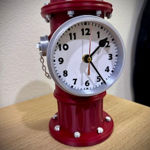 May include: A novelty clock designed as a red fire hydrant. The clock face is white with black numerals and black hands. The clock has silver-colored details and a small chain. The clock is approximately 20 cm tall.
