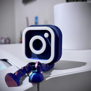 Op de afbeelding: Een 3D-geprint Instagram-logo in blauw en wit, met gelede poten en een kleine, bijpassende blauwe basis. Het logo is een vierkant met een witte rand en een wit camera-pictogram in het midden. De poten en basis hebben een metallic glans.
