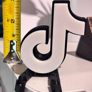 Peut inclure: Figurine du logo TikTok blanc et noir avec des jambes noires articulées. La figurine mesure environ 7,6 cm de haut, comme mesuré par un mètre ruban jaune en arrière-plan. Le logo est posé sur une surface blanche.