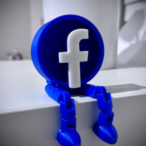 May include: A blue and white Facebook logo figure with articulated legs. The figure is sitting on a white surface. The Facebook logo is a white "f" on a blue circle.