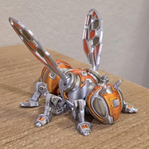 Op de afbeelding: Een gedetailleerd, 3D-geprint mechanisch insectenmodel. Het insect heeft een zilveren lichaam met oranje en rode accenten, inclusief de kop, vleugels en pootdetails. De vleugels hebben een honingraatpatroon. Het model staat op een houten oppervlak.