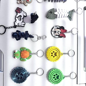Peut inclure: Une collection de porte-clés avec divers motifs, dont un bonhomme en pain d'épices, des squelettes de poissons, un lapin de dessin animé, une tortue et des empreintes de pattes. Les porte-clés sont en différents matériaux et ont des anneaux argentés.