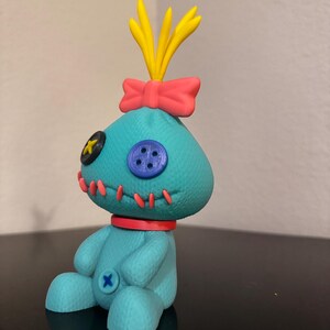 K&ouml;nnte beinhalten: T&uuml;rkisfarbene Pl&uuml;schpuppe mit aufgesticktem Design, rotem Kragen, rosa gen&auml;htem Mund und Schleife. Die Puppe hat gelbes Haar und eine rosa Schleife auf dem Kopf. Sie hat einen blauen Knopf und einen schwarzen Knopf im Gesicht.
