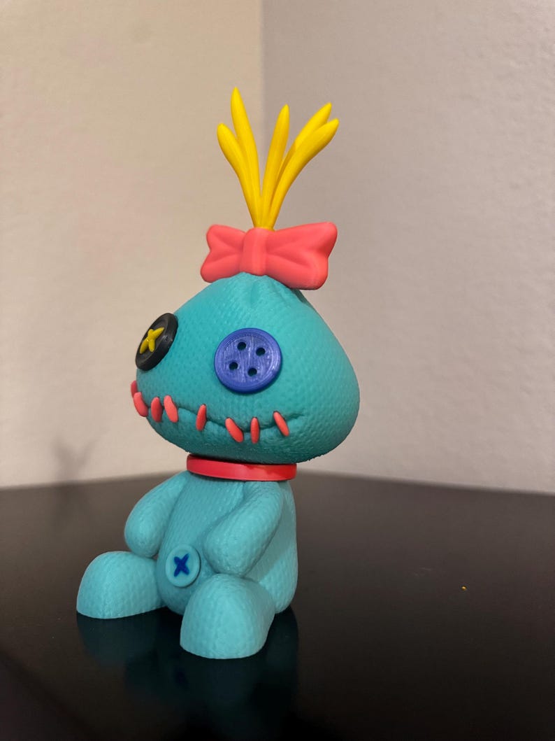 K&ouml;nnte beinhalten: T&uuml;rkisfarbene Pl&uuml;schpuppe mit aufgesticktem Design, rotem Kragen, rosa gen&auml;htem Mund und Schleife. Die Puppe hat gelbes Haar und eine rosa Schleife auf dem Kopf. Sie hat einen blauen Knopf und einen schwarzen Knopf im Gesicht.