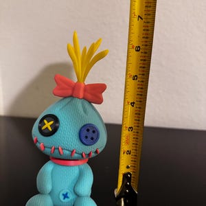 K&ouml;nnte beinhalten: T&uuml;rkisfarbene Pl&uuml;schpuppe mit Knopfaugen, einer roten Schleife und gelben haar&auml;hnlichen Akzenten. Die Puppe hat ein gen&auml;htes Aussehen mit rotem Faden und ist etwa 15 cm gro&szlig;. Ein gelbes Ma&szlig;band befindet sich neben der Puppe.