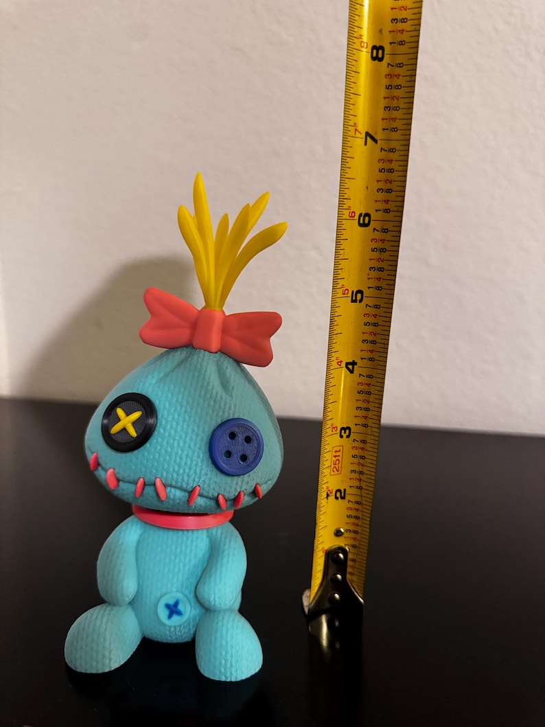 K&ouml;nnte beinhalten: T&uuml;rkisfarbene Pl&uuml;schpuppe mit Knopfaugen, einer roten Schleife und gelben haar&auml;hnlichen Akzenten. Die Puppe hat ein gen&auml;htes Aussehen mit rotem Faden und ist etwa 15 cm gro&szlig;. Ein gelbes Ma&szlig;band befindet sich neben der Puppe.