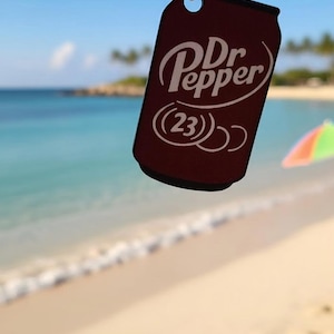 May include: A dark maroon, can-shaped pendant hanging from a silver-toned ball chain. The pendant features the white Dr Pepper logo with the number 23 in a circle below the text. The design is flat and mimics the silhouette of a soda can.