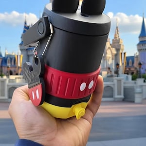 Könnte beinhalten: Ein schwarzer und roter Mickey-Mouse-Becher mit schwarzen Ohren oben. Der Becher hat ein rotes Band mit weißen Punkten und einen gelben Boden. Ein Mickey-Mouse-Schlüsselanhänger ist befestigt.