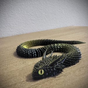 Puede incluir: Una escultura de dragón negro y verde con un cuerpo y una cabeza texturizados y detallados. El dragón tiene ojos amarillos y está posado en una posición curva. La escultura está sobre una superficie de madera.